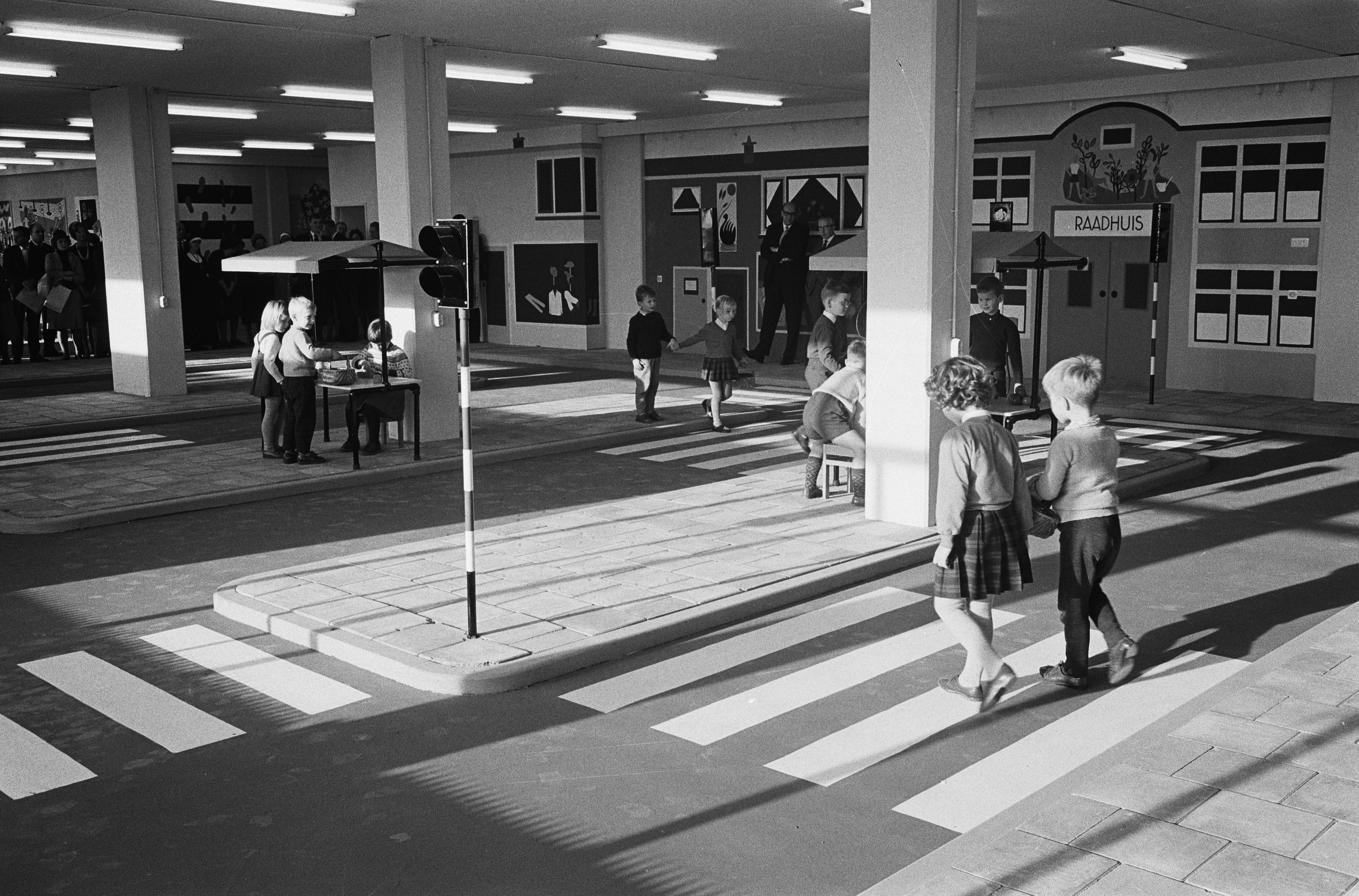Schwarz-weißes Foto von Kindern, die eine Straße an einer Ampel überqueren, mit Menschen auf Bänken und Säulen mit Lampen im Hintergrund.