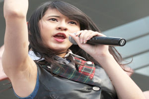 Eine Frau in einem weißen Hemd mit einem Pferdeschwanz singt leidenschaftlich in ein Mikrofon auf der Bühne, die Augen geschlossen, mit einem Gebäude im Hintergrund.