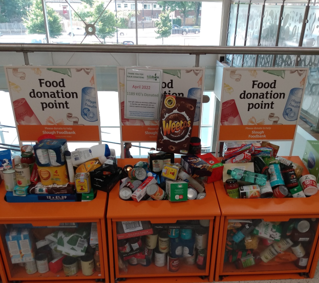 Orange Schübe in einem Gebäude voller gespendeter Lebensmittel wie Flaschen, Dosen und Kisten, mit Informationsschildern und einem Blick auf Fahrzeuge, Bäume, Gebäude und Himmel durch die Glasfenster.