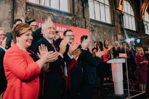 Eine Gruppe von Menschen, die vor einem Publikum stehen und applaudieren, mit einem Podium, einem Mikrofon und einer Tafel auf der rechten Seite und Stühlen, einer Fahne, einer Wand, Fenstern und Lichtern im Hintergrund.