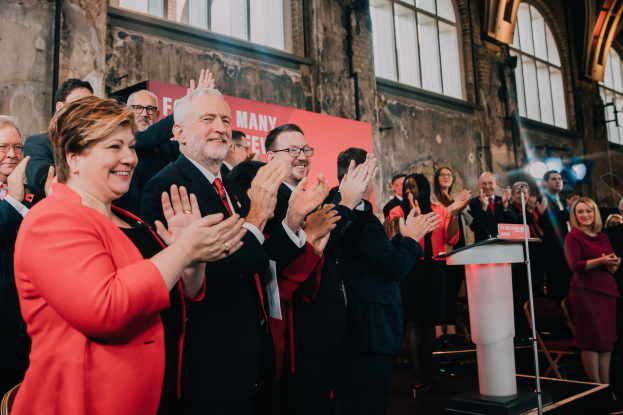 Eine Gruppe von Menschen, die vor einem Publikum stehen und applaudieren, mit einem Podium, einem Mikrofon und einer Tafel auf der rechten Seite und Stühlen, einer Fahne, einer Wand, Fenstern und Lichtern im Hintergrund.