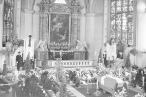 Schwarzes und weißes Foto einer Trauerfeier in einer Kirche mit Sitzenden und Stehenden, Blumensträußen, Kerzenständern und einem Sarg im Vordergrund, einem gerahmten Foto und Skulpturen an der Wand und Fenstern auf beiden Seiten.