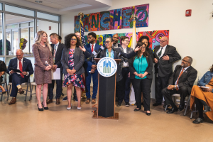 Gruppe von Menschen vor einem Rednerpult mit Mikrofon und einer Tafel mit der Aufschrift "Sancho Stadtrat tritt dem Stadtrat bei", mit sitzenden und stehenden Zuschauern, Flaggen, Feuerlöscher und Deckenleuchten im Hintergrund.