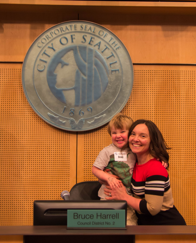 Eine Frau in einem roten und schwarzen Kleid hält ein Kind vor einem Podium mit Namensschild und Mikrofon, das vor einer Holzwand mit Logo steht.