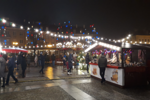Ein belebter Weihnachtsmarkt bei Nacht in einer Stadt, mit Menschen, die gehen, Ständen mit Lichtern und Dekorationen, Gebäuden mit Fenstern, Bäumen und festlichen Lichtern am Himmel.