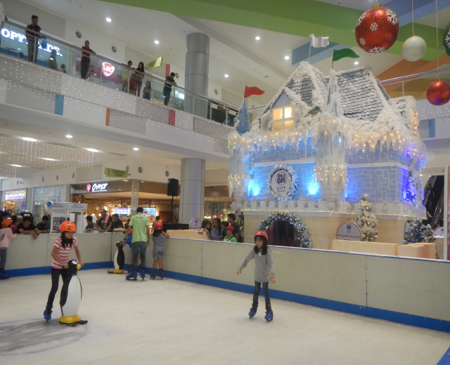 Eine Gruppe von Menschen, die auf einer Eisbahn in einem Einkaufszentrum Schlittschuh laufen, umgeben von einem hausähnlichen Aufbau, der mit Lichtern und festlichen Dekorationen geschmückt ist, mit Menschen, die auf dem Boden, Geländern und Säulen stehen, und Tafeln mit Text und Lichtern an der Decke.