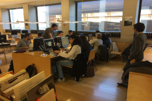 Gruppe von Menschen, die an Tischen in einer Bibliothek arbeiten, umgeben von Bücherregalen, Monitoren, Tastaturen, Mäusen, Taschen, Deckenleuchten und Glasfenstern.