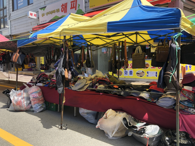 Ein Straßenmarkt mit Zelten, die Taschen, Hüte und andere Gegenstände ausstellen, vor einem Hintergrund aus Gebäuden mit Fenstern und Texttafeln.