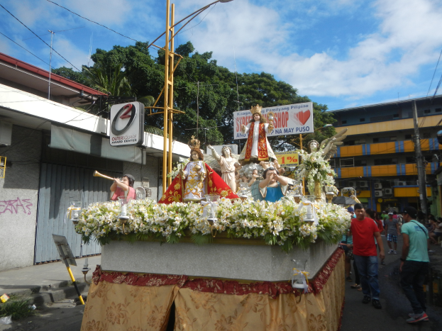 Ein Paradewagen mit Statuen, Blumen und Texttafeln, der eine Straße mit Laternenmasten, Drähten, Gebäuden, Bäumen und einem bewölkten Himmel entlangfährt.