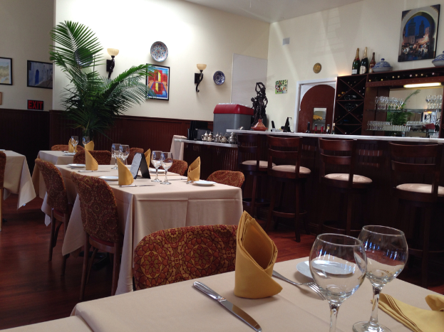 Ein Restaurantinnenraum mit Tischen und Stühlen, ein langer Tisch mit Gläsern, Servietten, Tellern und Löffeln, ein Regal mit Flaschen und Gläsern, eine Wand mit Foto Rahmen, eine Uhr, zwei Lampen und eine Pflanze vor der Wand.