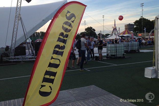 Ein buntes Volksfest-Szenario mit Zelten, einem zentralen Mast, Menschen, Bäumen, Strommasten, Straßenbahnen auf der Straße und einem eisernen Gestell links.