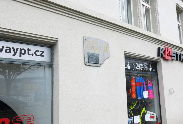 Ein Gebäude mit einem "Waypt.cz"-Schild, einer Glastür mit darauf hängenden Kleidungsstücken und einer Tafel an der Wand, die den Eingang zu einem Fahrradgeschäft in Prag anzeigt.