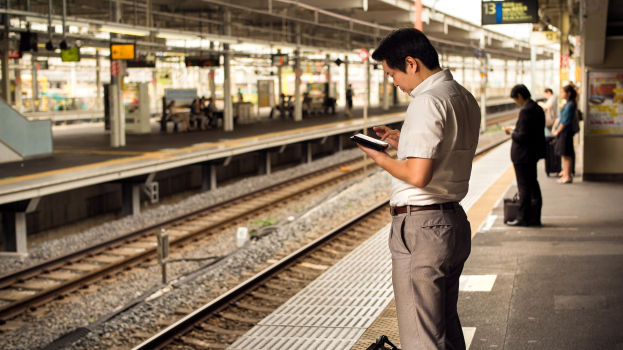 Ein Mann steht auf einem Bahnsteig und schaut auf sein Handy, umgeben von anderen Menschen mit Bahnschienen im Hintergrund; ein Schild mit Text ist auf der rechten Seite zu sehen.
