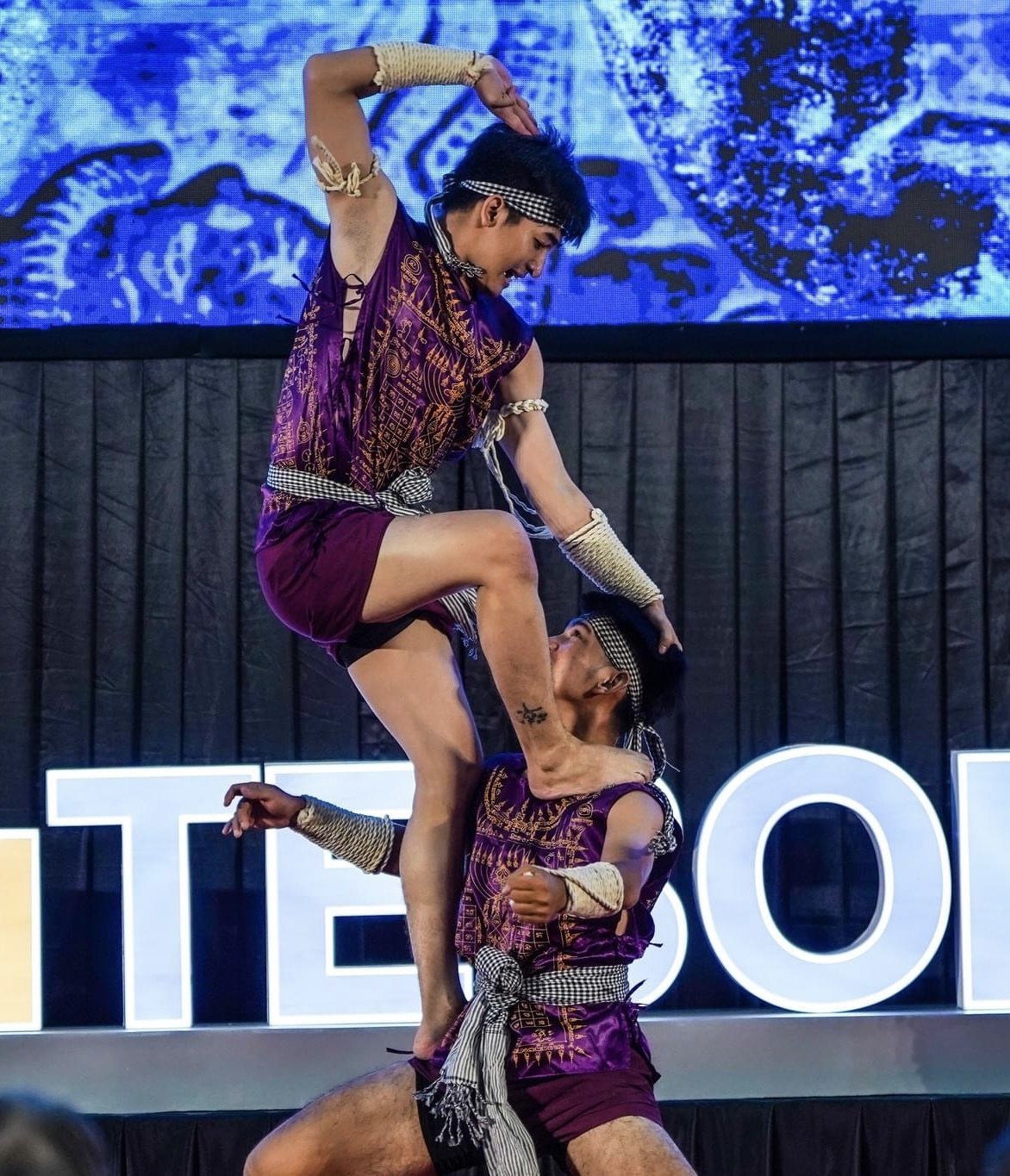 Zwei Performer in purpurfarbenen Kleidern führen einen akrobatischen Hebemanöver auf der Bühne vor einem Publikum aus, mit einem schwarzen Vorhang und einem Bildschirm im Hintergrund.
