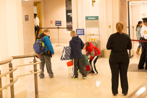 Eine Gruppe von Menschen steht in einem Flur neben einem Geländer und Deckenleuchten, mit Texttafeln im Hintergrund, die den ersten Schultag im U.S. Capitol suggerieren.
