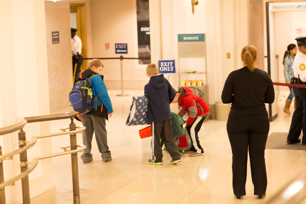 Eine Gruppe von Menschen steht in einem Flur neben einem Geländer und Deckenleuchten, mit Texttafeln im Hintergrund, die den ersten Schultag im U.S. Capitol suggerieren.