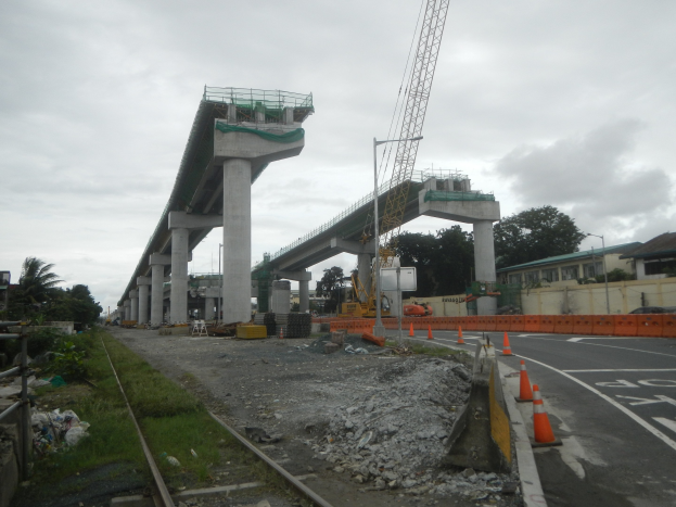 Baustelle mit einer Brücke im Hintergrund, eine Straße mit Absperrkegeln auf der rechten Seite, Steine und Gras auf dem Boden, eine Bahnschiene auf der linken Seite, Bäume und Gebäude auf beiden Seiten und ein bewölkter Himmel.