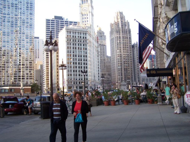Eine belebte Straßenszene mit Fußgängern, Autos und Blumenkübeln auf der Straße, mit Gebäuden, Straßenlaternen, Geschäften und Fahnen im Hintergrund.