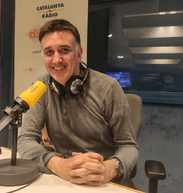 Ein Mann, der als Moderator von Catalunya Radio identifiziert wird, sitzt an einem Schreibtisch vor einem Mikrofon, trägt ein graues T-Shirt und einen Headset, mit einem Mikrofonständer, einem Kabel, einer Werbetafel und einem Glasfenster im Hintergrund.