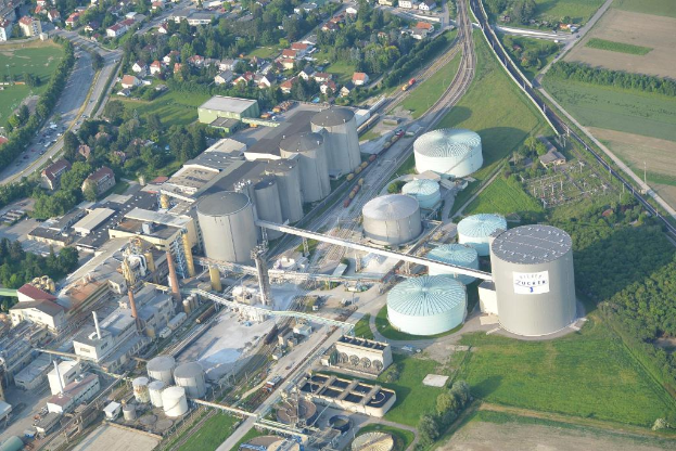 Luftaufnahme mit mehreren Gebäuden, Häusern, Bäumen und Fahrzeugen auf einer von Gras umgebenen Straße.