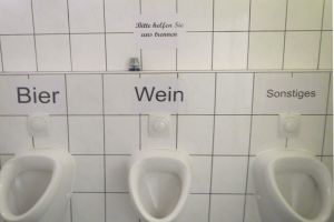 Drei weiße Toiletten an einer Wand montiert mit Schildern darüber, die "Bier Wein" und "Sonstiges" lesen.
