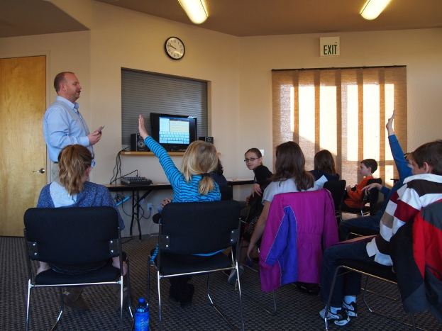 Ein Mann steht in einem Klassenzimmer und spricht zu einer Gruppe sitzender Kinder, mit einem Monitor, Lautsprechern, einer Uhr und einem Fenster dahinter und einer Tür links daneben.