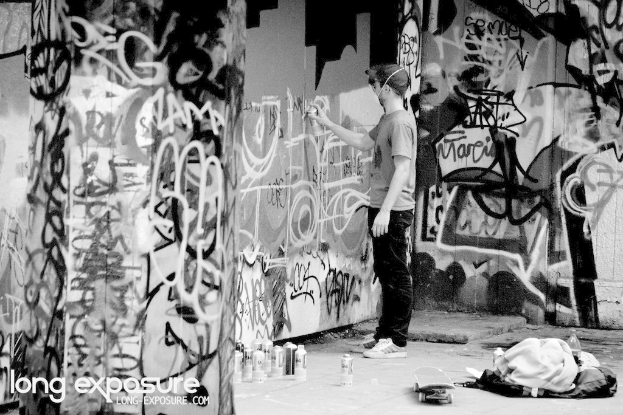 Ein Mann steht drinnen und h√§lt eine Flasche in der Hand, mit verstreuten Gegenst√§nden wie anderen Flaschen, einem Skateboard, einer Wasserflasche und Klamotten auf dem Boden, Graffiti an den W√§nden und Text unten links.