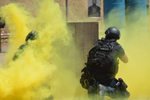 Zwei Polizisten in Einsatzausrüstung mit gelbem Rauch aus dem Mund, die Helme, Taschen und andere Schutzausrüstung tragen, stehen vor einer Wand mit einer Tafel mit Text darauf und anderen Gegenständen, wobei der Boden unten sichtbar ist.