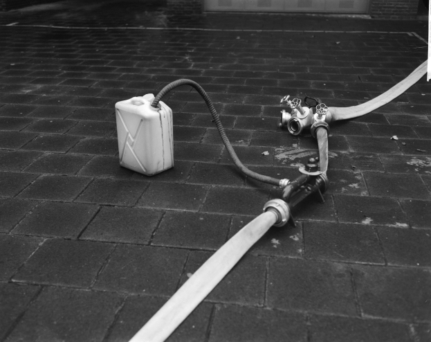 Schwarz-weißes Foto eines Gasbehälters mit einem Schlauch, der vor einem Gebäude mit einer Tür im Hintergrund auf dem Boden liegt.