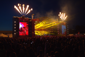 Eine große Menschenmenge auf einem Musikfestival mit einer Bühne in der Mitte, Musiker, die unter hellen Lichtern spielen, Banner mit Text, Zelte links und ein dunkler Himmel mit Feuerwerk im Hintergrund.