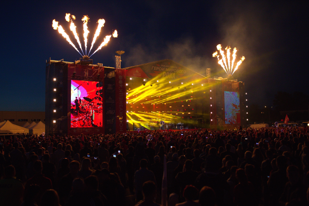 Eine große Menschenmenge auf einem Musikfestival mit einer Bühne in der Mitte, Musiker, die unter hellen Lichtern spielen, Banner mit Text, Zelte links und ein dunkler Himmel mit Feuerwerk im Hintergrund.