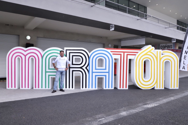 Ein Mann steht am Start eines Marathons vor einem großen "Marathon"-Schild, mit einer Fahne rechts und einem Gebäude mit Lichtern, Geländern und Läden im Hintergrund.