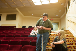 Ein Mann in einem grünen T-Shirt und blauen Jeans steht in einem Theater und hält ein Mikrofon in der Hand, hinter ihm sitzen zwei Frauen auf einer Treppe, rote Stühle im Hintergrund, ein Geländer auf der rechten Seite und Lampen auf dem Dach.