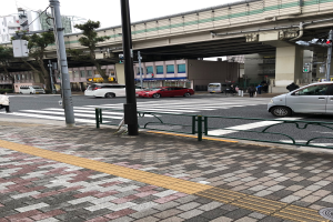 Stadtstraße mit Fahrzeugen, Fußgängern an einer Ampel, Verkehrszeichen, einer Brücke, Bäumen, Gebäuden und einem klaren blauen Himmel.
