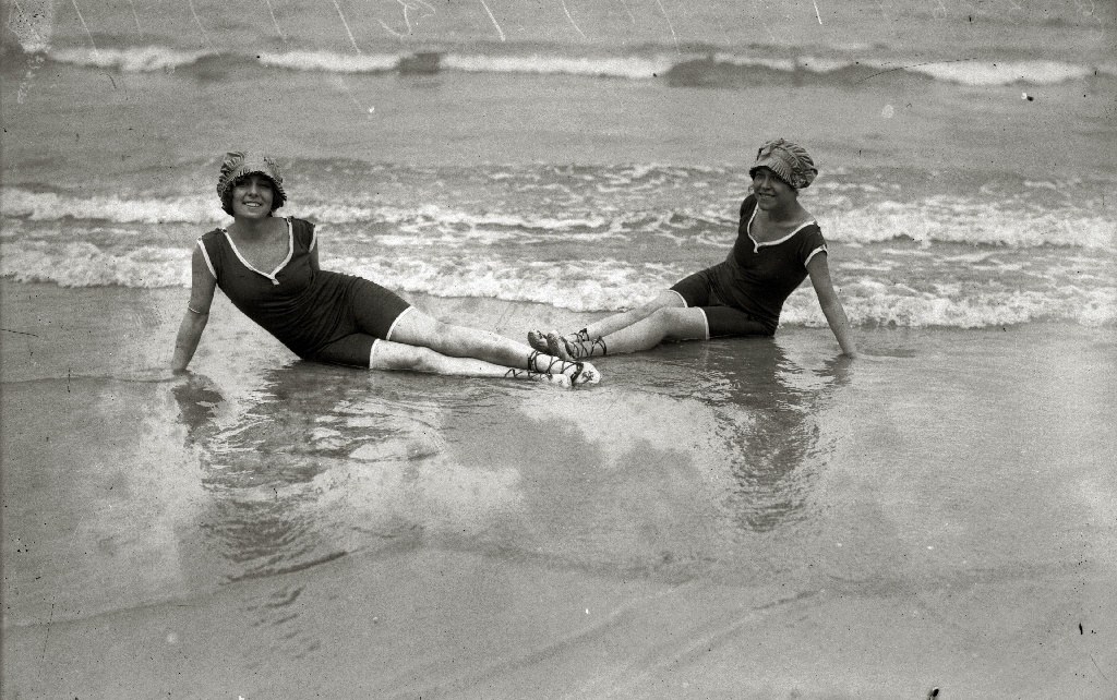 Zwei Frauen in Bikinis und Hüten sitzen im Wasser am Strand, lächeln und genießen die Sonne und die Wellen, in schwarz-weiß.