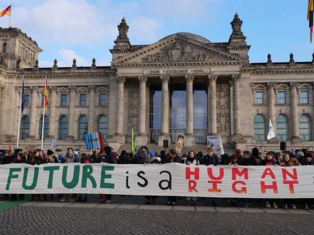 Eine Gruppe von Menschen mit einem Transparent "Zukunft ist ein Menschenrecht" vor dem Reichstaggebäude in Berlin, Deutschland, mit sichtbaren Fenstern, Säulen und Bögen sowie umgeben von Fahnen mit Stangen unter einem bewölkten Himmel.