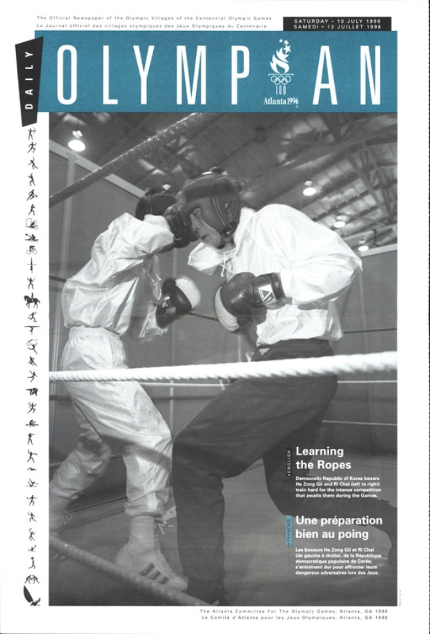 Zwei Boxer in einem Boxring, die Helme und Handschuhe tragen, mit Seilen um den Ring und beleuchteter Szene, mit 'Olympian Magazine'-Text oben und unten.
