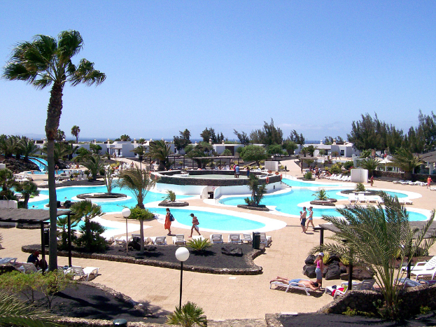 Ein Resort mit Pools umgeben von Bäumen, Menschen zwischen den Pools, Gebäuden im Hintergrund und einem klaren Himmel überall.