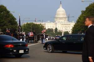 Eine Trauerprozession mit Menschen in Mützen und mit Fahnen zieht am Capitol Building vorbei, begleitet von Fahrzeugen, Laternenmasten, Bäumen und Gebäuden unter einem klaren Himmel.