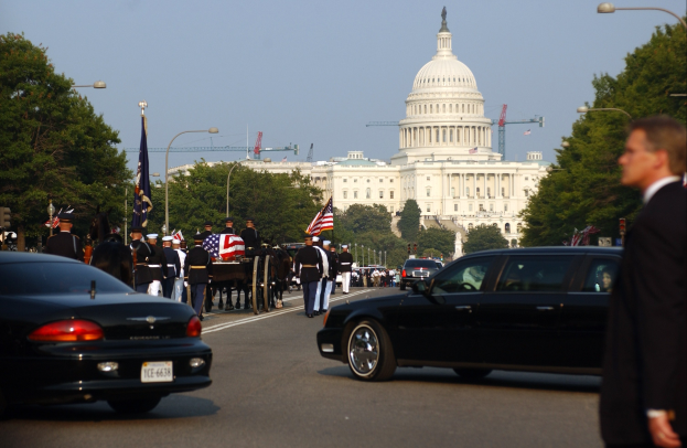 Eine Trauerprozession mit Menschen in Mützen und mit Fahnen zieht am Capitol Building vorbei, begleitet von Fahrzeugen, Laternenmasten, Bäumen und Gebäuden unter einem klaren Himmel.