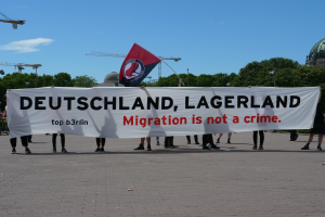 Eine Gruppe von Menschen, die draußen stehen und ein Banner halten, auf dem "Deutschland, Lagerland Migration ist kein Verbrechen" und eine Flagge zu sehen ist, mit Straßenlaternen, Bäumen, Kränen, einem Gebäude und einem bewölkten Himmel im Hintergrund.