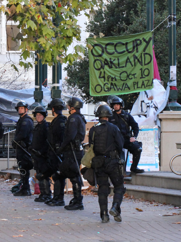Eine Gruppe von Polizisten in Helmen, die Waffen halten, geht eine von Bäumen gesäumte Straße mit verschiedenen Gegenständen und einem Gebäude im Hintergrund entlang.