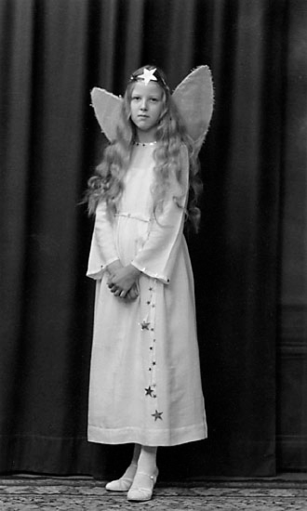 Ein junges Mädchen in einem weißen Kleid mit Engelsflügeln steht auf einer Bühne vor einem Vorhang, trägt Schuhe und ist bereit für eine Vorstellung, dargestellt in Schwarz-Weiß.