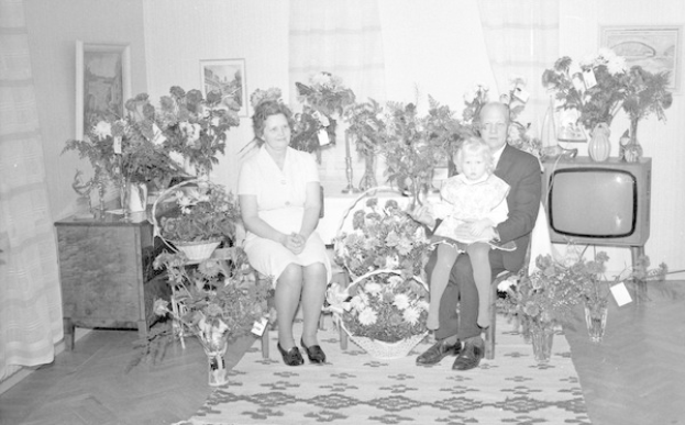 Schwarzes und weißes Foto einer Familie aus drei Personen - einem Mann, einer Frau und einem Kind - auf einem Sofa sitzend vor einem Fernseher, Blumenvasen auf den Seitentischen, ein Teppich auf dem Boden, Vorhänge und Fotoalben an der Wand.