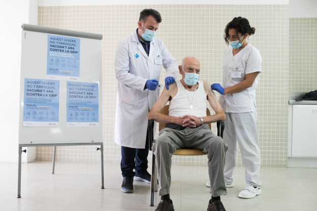 Ein älterer Mann sitzt in einem Stuhl und erhält von zwei maskierten und handschuhgetragenen Ärzten eine Impfung in einer Krankenhausumgebung, mit einer Tafel links und Gegenständen auf einer Plattform rechts.
