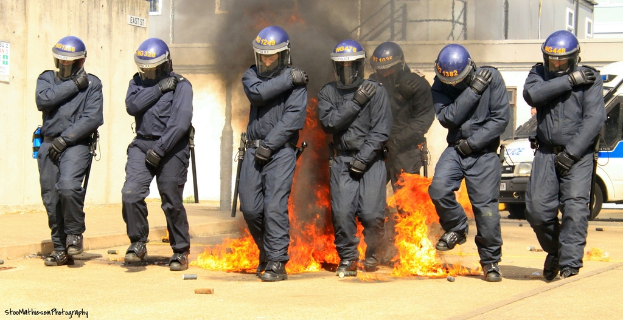 Menschen in Helmen stehen vor einem Feuer mit verschiedenen Gegenständen auf dem Boden, Gebäuden im Hintergrund, einem Fahrzeug und einem Plakat und einer Tafel an der linken Wand; Text ist unten im Bild sichtbar.