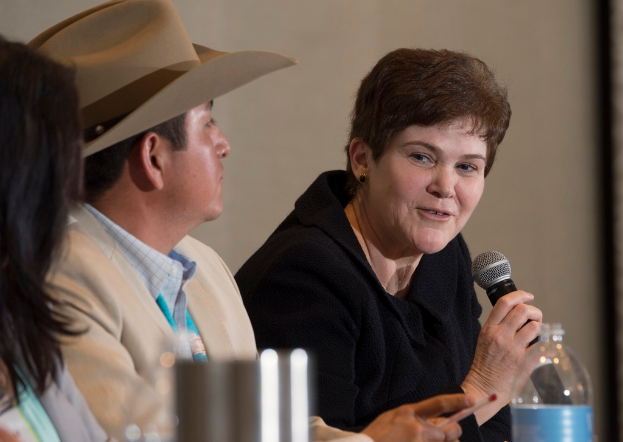 Drei Personen sitzen in einem Raum, eine Frau in einer schwarzen Jacke hält ein Mikrofon, ein Mann in einem cremefarbenen Blazer trägt eine Mütze und hat eine Wasserflasche vor sich und eine weiße Wand hinter sich.