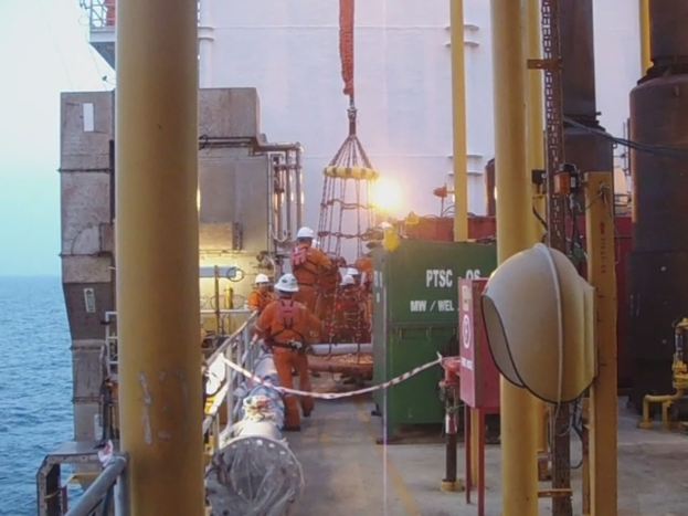 Ein Schiff mit Masten, Kisten und Geländern, mit Menschen in Helmen vor Wasser.