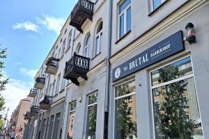Brutalistischer Barbershop-Laden in Berlin mit Betongebäuden, Fenstern, Geländern, Balkonen und Beschriftung, umgeben von Bäumen, Straßenlaternen und Fußgängern unter einem bewölkten Himmel.
