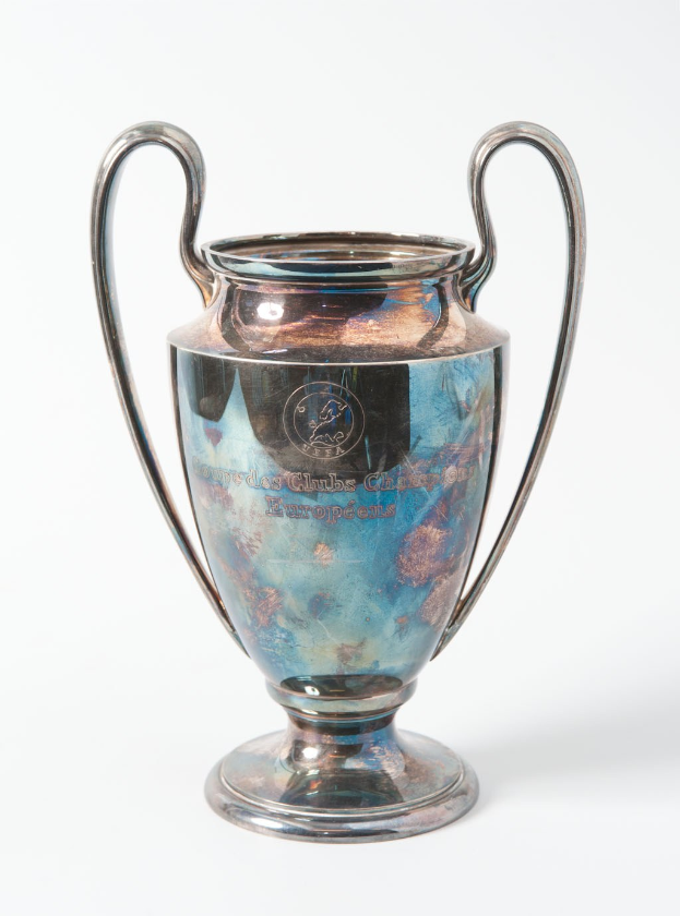 Der UEFA-Champions-League-Pokal, ein stählerner Becher mit zwei Griffen und einem blauen und silbernen Farbschema, der mit den Worten 'UEFA Champions League' auf einem weißen Hintergrund eingraviert ist.